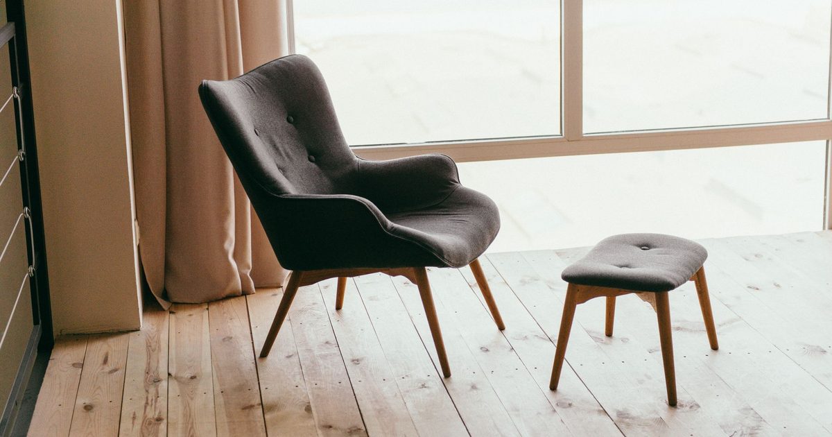 a cushioned footstool beside a grey armchair by a bright window