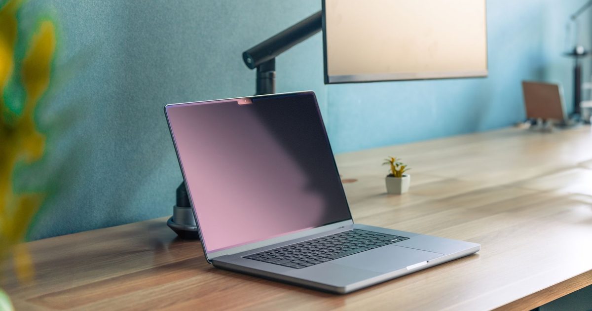 a laptop on a wooden desk with a monitor on an arm behind it