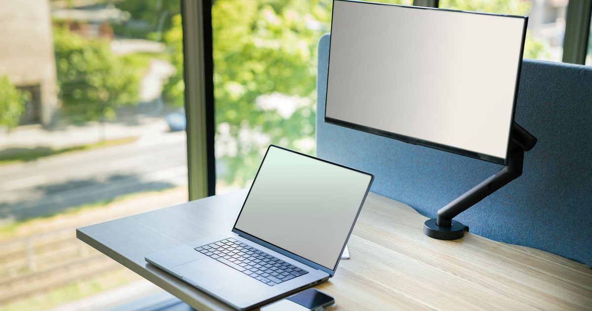 a monitor held on a black adjustable arm beside a laptop by a window
